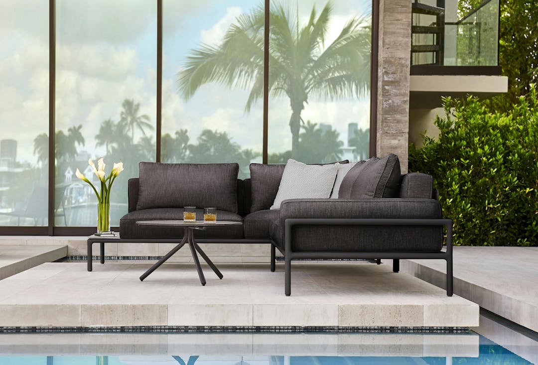 Ann Marie Vering's Moto collection, modular edition in black finish and black fabric with black and grey pillows set up outside a beautiiful home with a contemporary pool. The modular sofa is L-shaped.