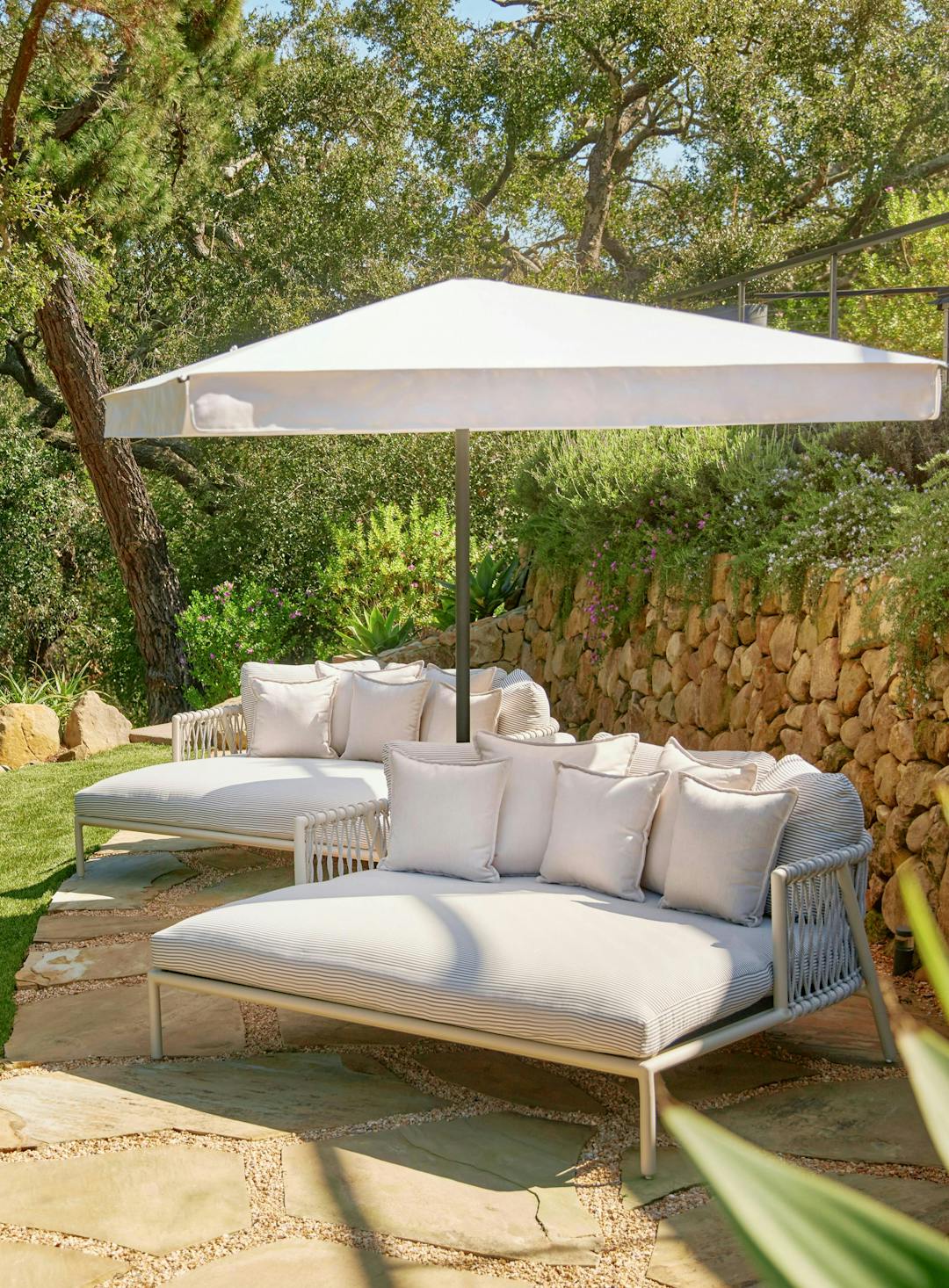 2 daybeds with white fabric and finish underneath white umbrella with stone wall behind. Oscar Daybeds by Ann Marie Vering.