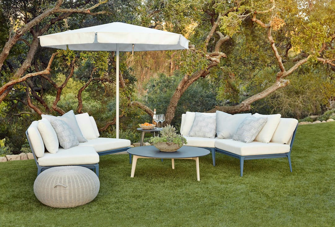 Still modular collection with white fabric, white and grey pattern pillows, and blue finish with round coffee table, side table, and umbrella on a bright green lawn and forest in the background. There's a round Juno ottoman on the side.
